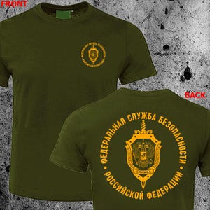 May include: Olive green t-shirt with a gold emblem on the front and back. The front emblem is a circular design with text. The back of the shirt features a larger circular emblem with text and a shield design.