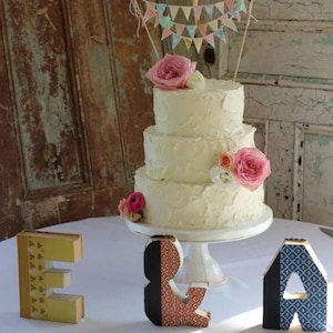 Puede incluir: Un pastel de tres pisos con glaseado blanco, adornado con rosas rosas y un adorno de pastel de banner. El pastel está sobre un pedestal blanco, con letras decorativas que deletrean "E & A" al frente. Las letras están decoradas con papel estampado.