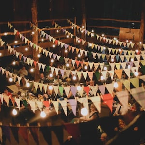 May include: A large group of people gathered around tables in a dimly lit room decorated with colorful pennant flags and string lights. The flags are strung across the ceiling and the lights are strung along the flags.