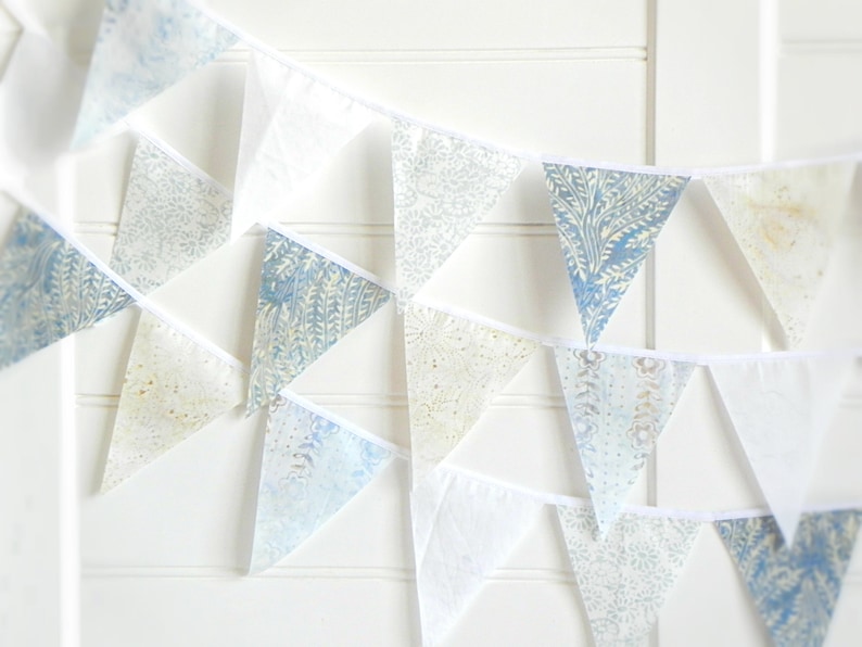 May include: A blue and white patterned bunting with triangular flags. The flags are made of a light fabric and have a white trim. The bunting is strung on a white cord.