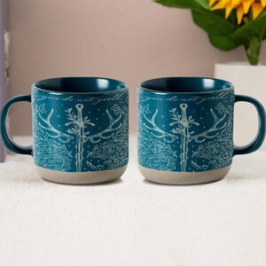 May include: Two teal ceramic mugs with a decorative design featuring a sword, antlers, and the phrase "you do not yield" in white. The mugs have a dark teal interior and a light beige base.
