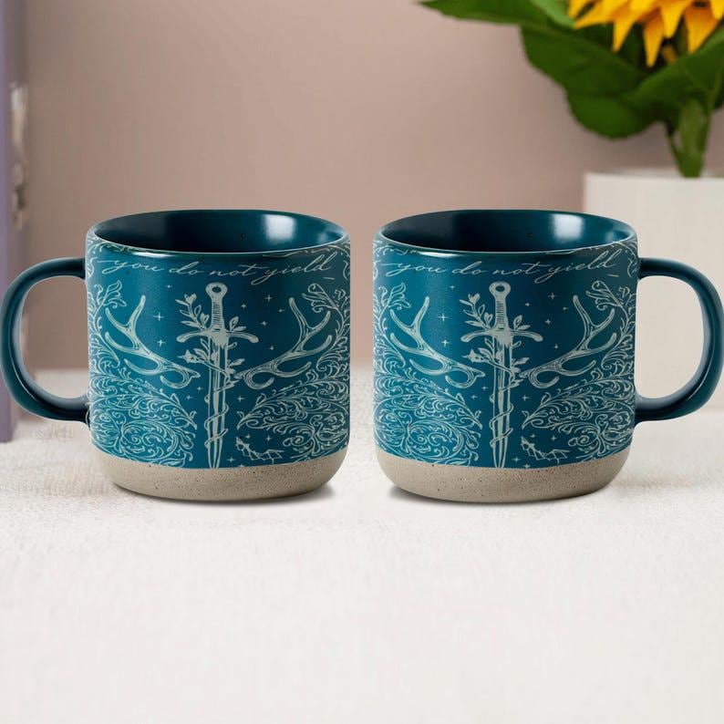 May include: Two teal ceramic mugs with a decorative design featuring a sword, antlers, and the phrase "you do not yield" in white. The mugs have a dark teal interior and a light beige base.