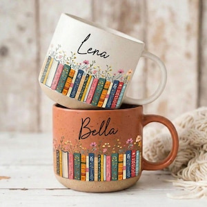 Puede incluir: Dos tazas de cerámica apiladas. La taza superior es blanca con el nombre "Lena" en escritura negra. La taza inferior es naranja con el nombre "Bella". Ambas tazas presentan un diseño colorido de estantería con detalles florales.