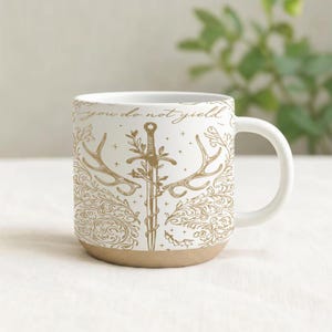 May include: White ceramic mug with a tan base and a curved handle. The mug features a gold illustration of a sword, antlers, and floral elements, with the text "you do not yield" in gold lettering. The mug is designed for hot beverages.