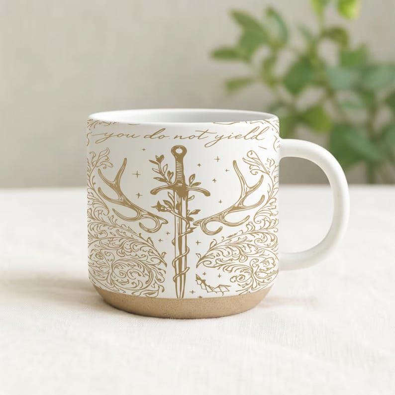 May include: White ceramic mug with a tan base and a curved handle. The mug features a gold illustration of a sword, antlers, and floral elements, with the text "you do not yield" in gold lettering. The mug is designed for hot beverages.