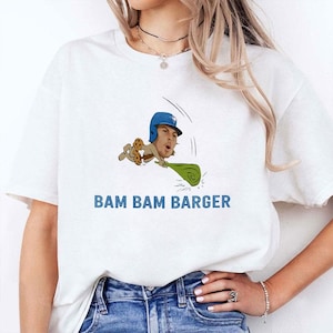 May include: White t-shirt with a cartoon baseball player illustration and the blue text "BAM BAM BARGER". The player wears a blue helmet and holds a green object. The design is centered on the shirt.