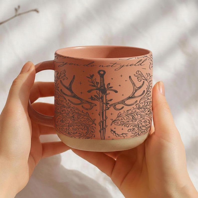 May include: A ceramic mug with a peach-colored exterior and a cream-colored base. The mug features a black illustration of a sword, antlers, and floral elements, with the words "you do not yield" in script. The mug is held by two hands.