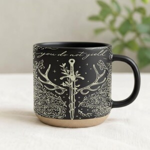 May include: Black ceramic mug with a cream-colored design featuring a sword, antlers, and floral elements. The text "you do not yield" is written around the top of the mug. The bottom of the mug is a natural tan color.