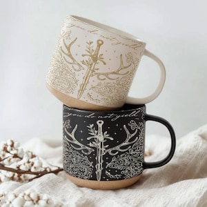 May include: Two ceramic mugs stacked on top of each other. The top mug is off-white with a gold design of a sword, antlers, and the words "you do not yield." The bottom mug is black with a similar design in white.