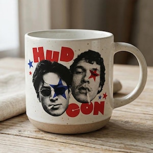 May include: A ceramic coffee mug with a cream-colored speckled finish and a tan base. The mug features two male faces with star-shaped eye designs and the word "HUDSON" in red. One face has a blue star, the other a red star.