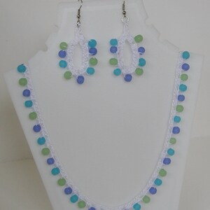 Op de afbeelding: Een witte gehaakte ketting en bijpassende oorbellen met blauwe, groene en witte kralen.