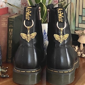 May include: A pair of black leather ankle boots featuring yellow stitching along the sole and a black heel pull tab with the yellow text AirWair. Each boot is adorned with a gold-toned metal moth charm attached to a ring on the heel tab.