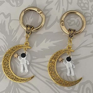 May include: Two gold-colored keychains with a crescent moon design. Each moon has an astronaut charm holding a star. The keychains have a decorative clasp and are suspended from a decorative connector.