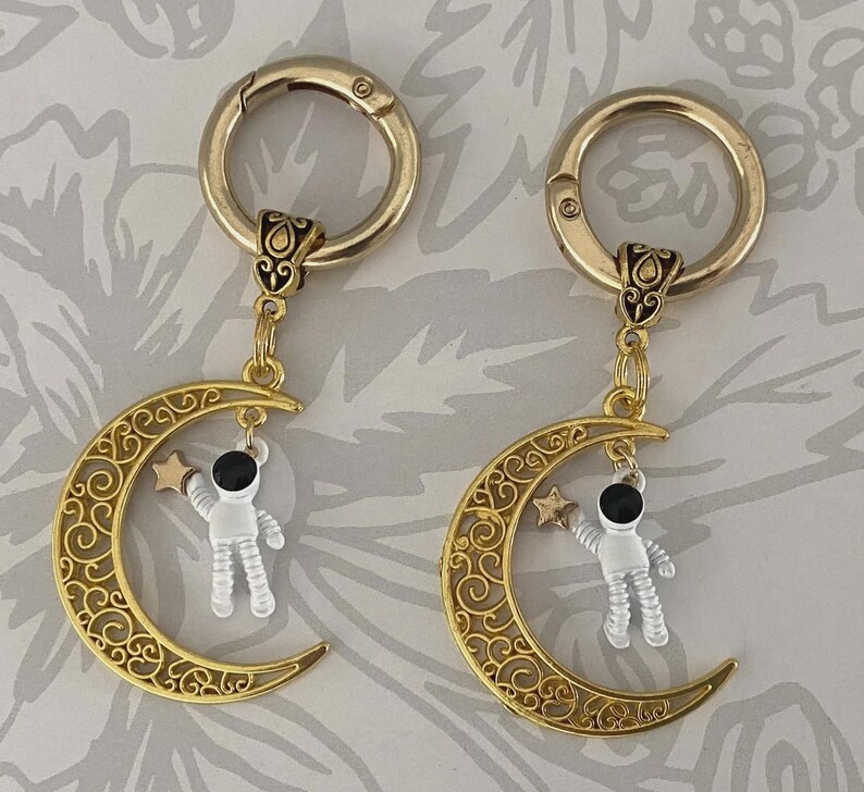 May include: Two gold-colored keychains with a crescent moon design. Each moon has an astronaut charm holding a star. The keychains have a decorative clasp and are suspended from a decorative connector.