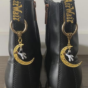 May include: Black leather boots with gold-colored charms. Each charm features a crescent moon design with a small astronaut figure. The boots have gold-colored zippers and pull tabs with the text "Air Wair" in gold.