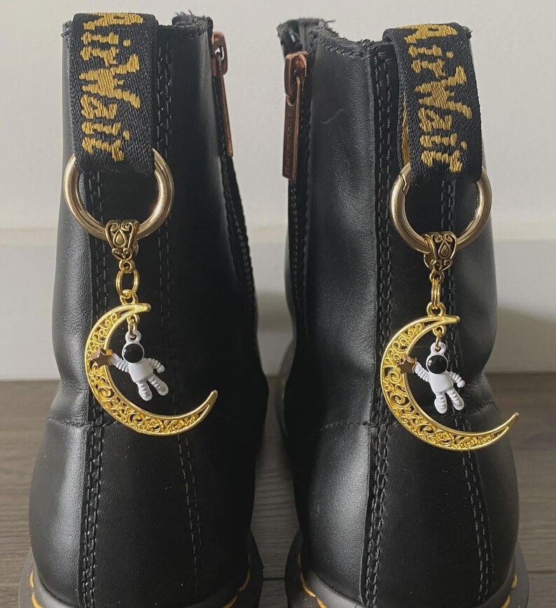 May include: Black leather boots with gold-colored charms. Each charm features a crescent moon design with a small astronaut figure. The boots have gold-colored zippers and pull tabs with the text "Air Wair" in gold.