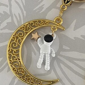May include: A gold-colored keychain featuring a crescent moon with intricate scrollwork. A small astronaut figure in white and black hangs from the moon, holding a gold star. The keychain has a gold ring and a decorative top.