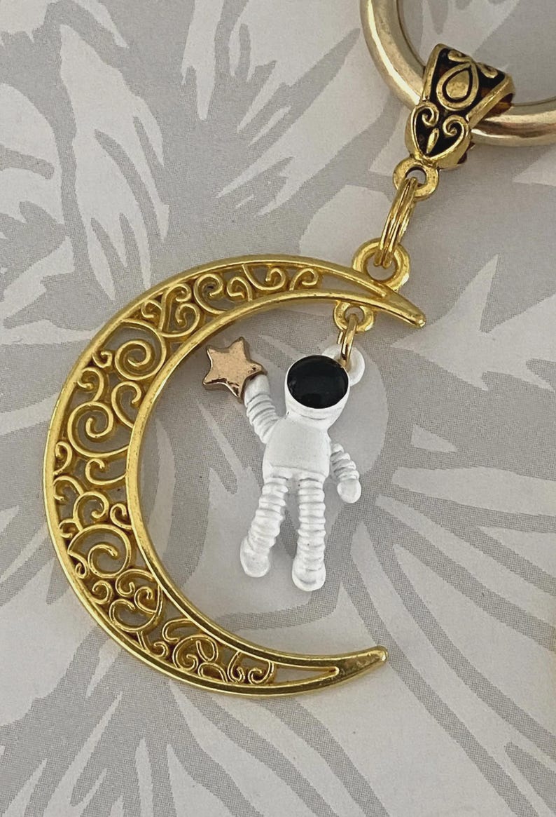 May include: A gold-colored keychain featuring a crescent moon with intricate scrollwork. A small astronaut figure in white and black hangs from the moon, holding a gold star. The keychain has a gold ring and a decorative top.