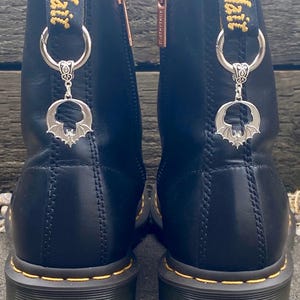 May include: Black leather boots with yellow stitching and a silver bat charm. The boots feature a zip and a pull tab with "Air Wair" in yellow text. The bat charm is suspended from a silver ring.