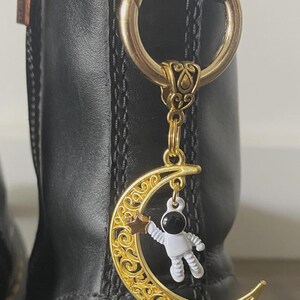 May include: A gold-colored boot charm featuring a crescent moon with intricate filigree and a small astronaut figure. The charm hangs from a gold ring attached to a black boot.