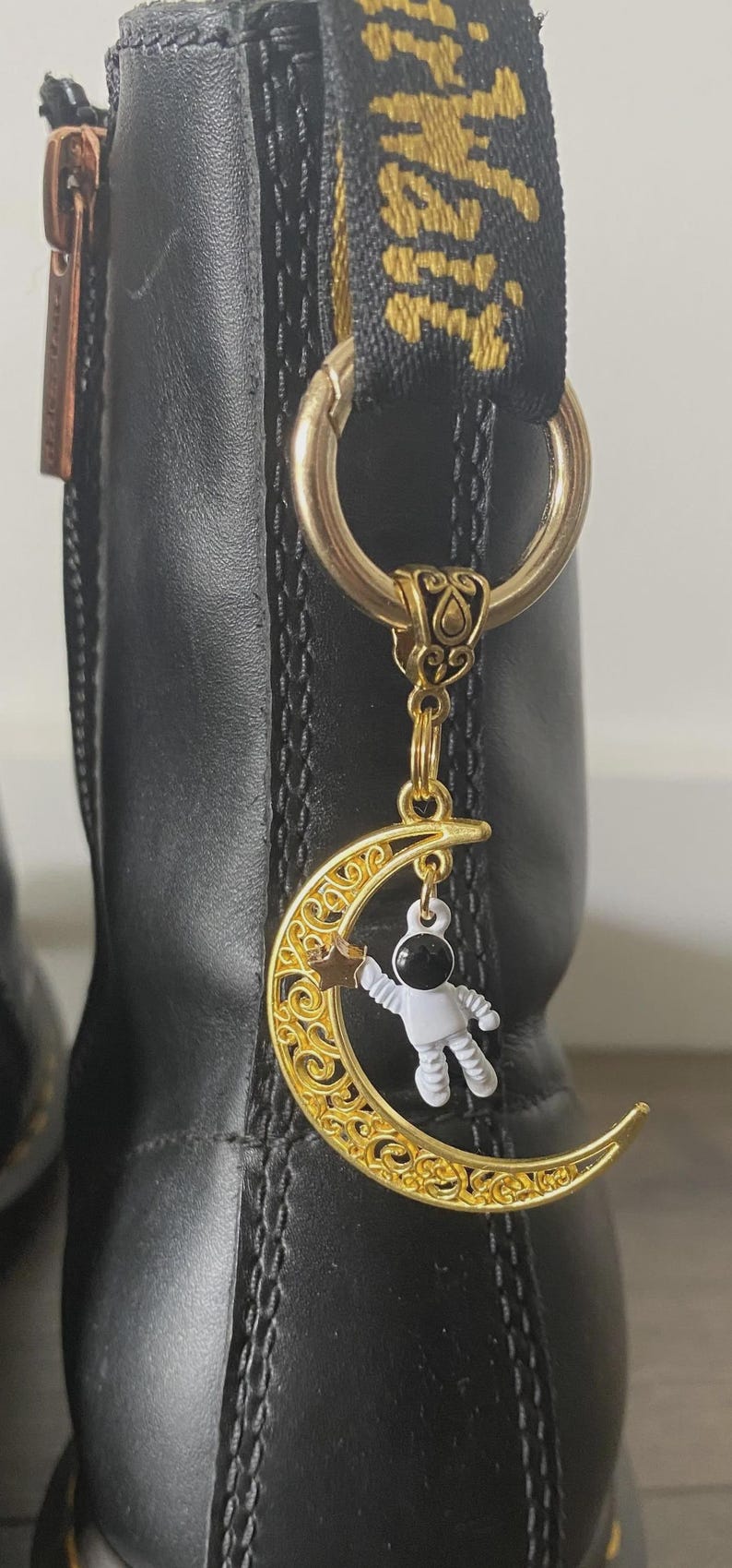 May include: A gold-colored boot charm featuring a crescent moon with intricate filigree and a small astronaut figure. The charm hangs from a gold ring attached to a black boot.