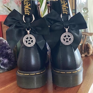 May include: A pair of black leather ankle boots featuring yellow stitching along the sole and a yellow AirWair heel loop. Each boot is adorned with a black satin bow attached to a silver-toned metal ring, which holds a circular pentacle charm.