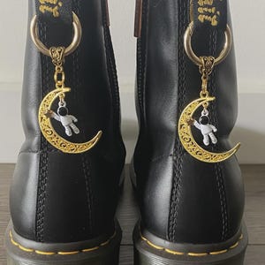 May include: Pair of black leather boots with gold-colored moon and astronaut charms hanging from the top. The boots have yellow stitching and a zipper. The charms are detailed with a gold crescent moon and a small astronaut figure.