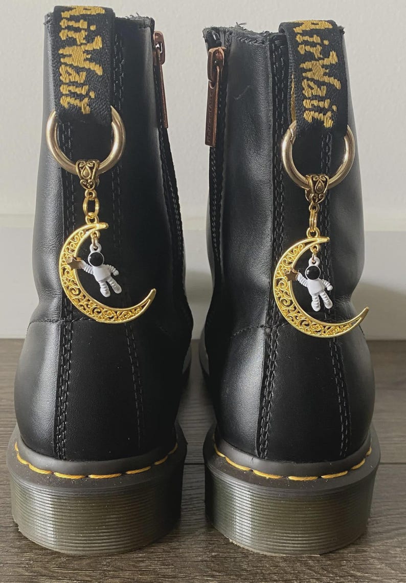 May include: Pair of black leather boots with gold-colored moon and astronaut charms hanging from the top. The boots have yellow stitching and a zipper. The charms are detailed with a gold crescent moon and a small astronaut figure.