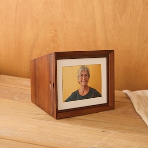 May include: A wooden photo frame with a portrait of a person. The frame is a dark brown color and has a square shape. The photo is displayed behind a white mat. The frame is sitting on a wooden surface.