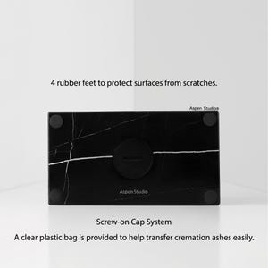 May include: Black marble cremation urn with white veining, featuring a screw-on cap system. The urn has four rubber feet to protect surfaces. The text "Aspen Studio" is visible. A clear plastic bag is provided to help transfer cremation ashes easily.