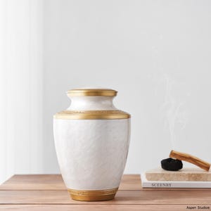 May include: A white urn with a textured surface and gold-colored accents. The urn has a lid and decorative bands around the top and base. The urn is on a wooden surface.