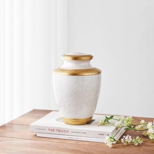 May include: A white urn with a textured surface and gold accents. The urn has a lid and decorative gold bands around the top and base. It sits on a stack of books with the title "THE TOUCH".