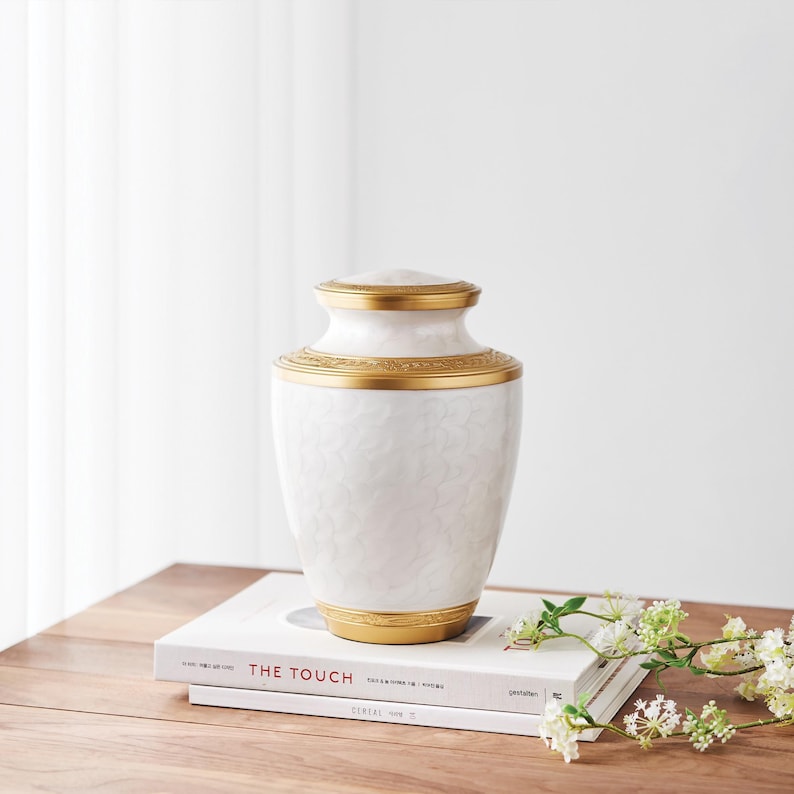 May include: A white urn with a textured surface and gold accents. The urn has a lid and decorative gold bands around the top and base. It sits on a stack of books with the title "THE TOUCH".