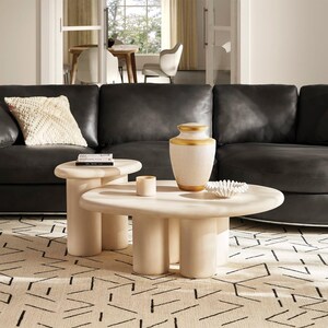 May include: A living room scene featuring a black leather sofa, a cream-colored rug with a black geometric pattern, and two cream-colored coffee tables. A decorative vase with gold accents sits on the larger table.