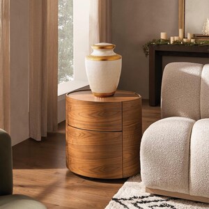 May include: A decorative white and gold urn sits atop a round, wooden nightstand with two drawers. The urn has a gold band around the top and bottom. The nightstand is made of wood with a visible grain pattern. The scene is set in a well-lit room with a neutral color palette.