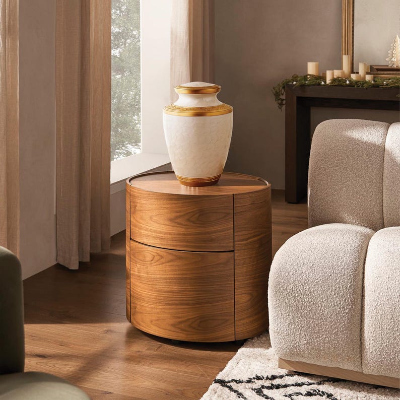May include: A decorative white and gold urn sits atop a round, wooden nightstand with two drawers. The urn has a gold band around the top and bottom. The nightstand is made of wood with a visible grain pattern. The scene is set in a well-lit room with a neutral color palette.