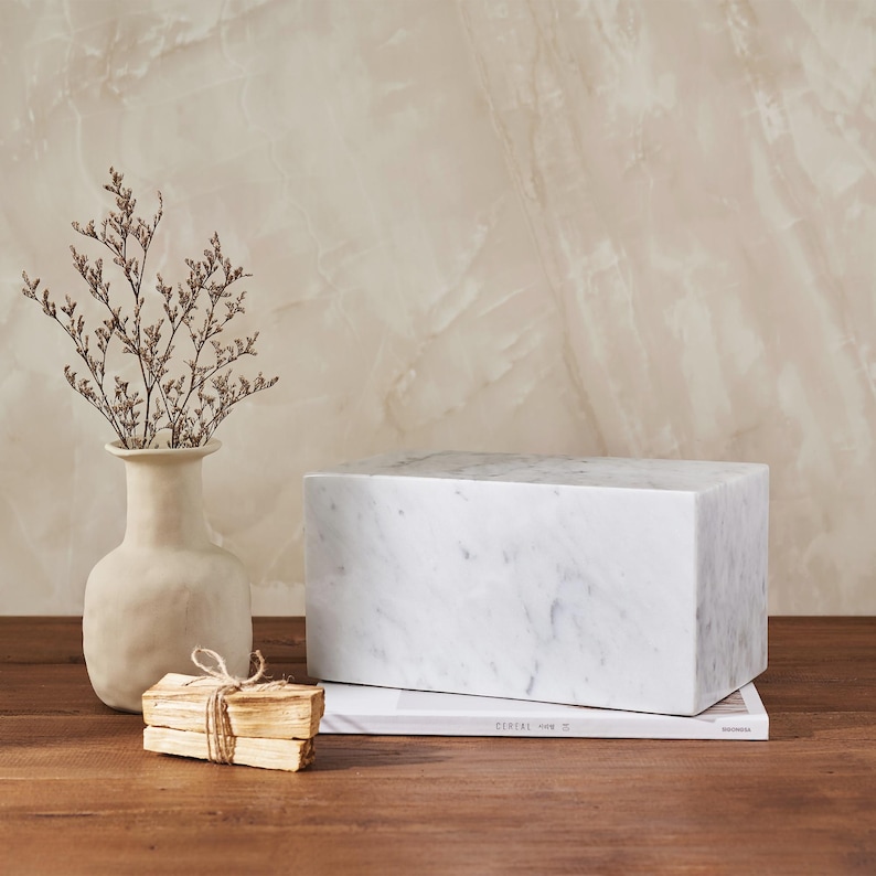May include: A white marble block sits on a wooden surface with a stack of wooden sticks and a vase with dried flowers. The marble block is a rectangular shape with a smooth surface. The wooden sticks are tied together with twine. The vase is a light brown color and has a rounded shape.