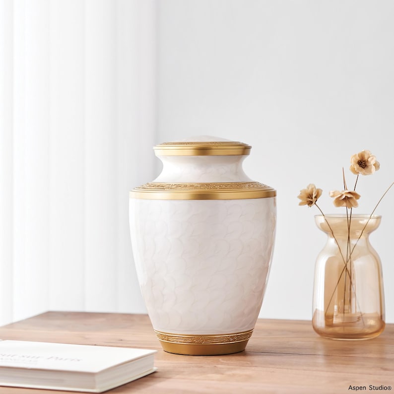 May include: A white urn with a gold rim and accents. The urn has a textured pattern and a matching lid. A small glass vase with dried flowers sits to the right of the urn. The urn is on a wooden surface.