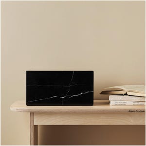 May include: A rectangular black marble block with white veining sits on a light wood table. The block is the focal point, with a neutral background and a stack of books visible on the right.