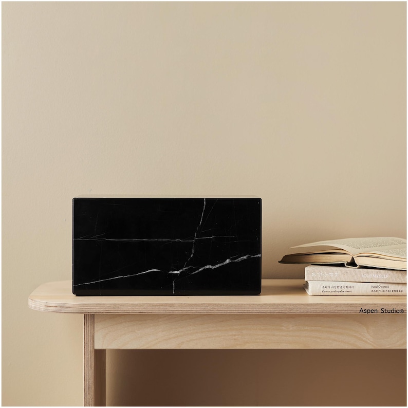 May include: A rectangular black marble block with white veining sits on a light wood table. The block is the focal point, with a neutral background and a stack of books visible on the right.