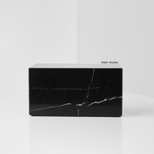 May include: A rectangular black marble block with white veining. The block is a solid, minimalist design, suitable for various uses. The surface is polished, reflecting light. The Aspen Studio logo is visible on the top right corner.