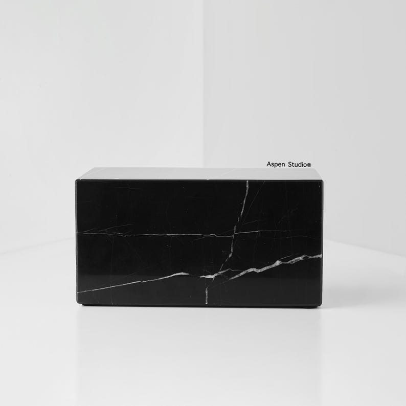 May include: A rectangular black marble block with white veining. The block is a solid, minimalist design, suitable for various uses. The surface is polished, reflecting light. The Aspen Studio logo is visible on the top right corner.