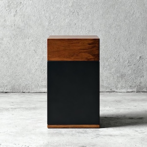May include: A rectangular wooden box with a dark brown top and bottom frame, and a black center. The box is sitting on a concrete surface. The wood has a natural grain.