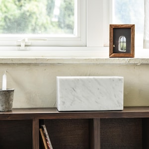 May include: A white marble speaker sits on a dark wood shelf with a candle and a potted plant. The shelf is part of a bookcase with other shelves and books.