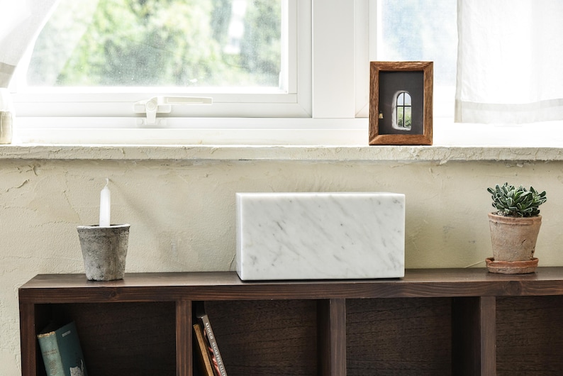 May include: A white marble speaker sits on a dark wood shelf with a candle and a potted plant. The shelf is part of a bookcase with other shelves and books.