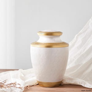 May include: A white urn with a gold-colored lid and base, and gold trim around the body. The urn has a subtle, repeating pattern on its surface. The urn is a decorative item.