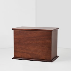 May include: A rectangular wooden box with a rich, dark brown finish. The box has a flat top with a slightly rounded edge and sits on a matching base. The wood grain is visible, adding to the natural aesthetic. The box is designed with clean lines and a simple, classic style.