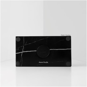May include: A rectangular black marble object with white veining, featuring a circular indentation and the text "Aspen Studio". The object has four small, round, black rubber feet.