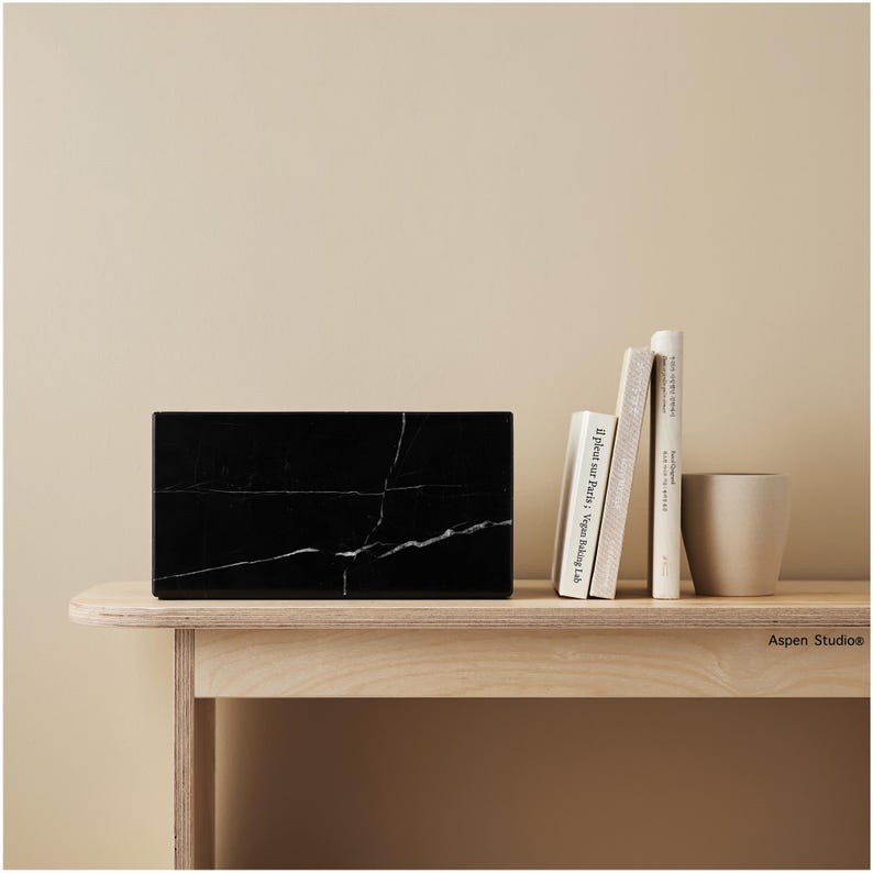 May include: A rectangular black marble box with white veining sits on a light wood table. Several books and a beige ceramic cup are arranged nearby. The background is a solid, neutral color.