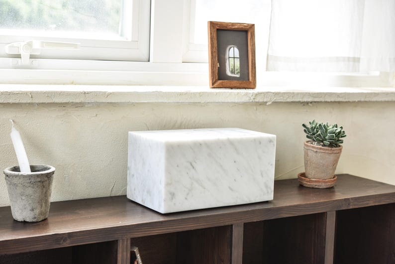 May include: A white marble box with a smooth, polished surface sits on a dark wood shelf. A small potted succulent plant is on the shelf to the right of the box.